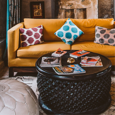 A warm and cozy living room with deep yellow couch, three throw pillows and a dark wood coffee table which has intricate criss-cross carving on the side.